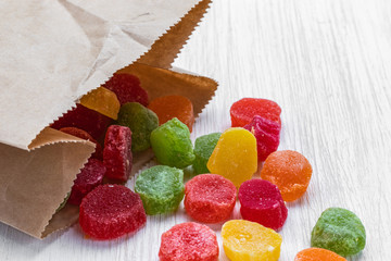 Multicolored round slices of dried pineapple in sugar lie in a paper bag and on a white wooden surface, side view from above