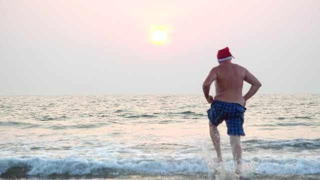 A fat man in a red Santa hat runs funny holding shorts and dives into the sea at sunset. A goofy Christmas image of a running fat Santa Claus in the sea.
