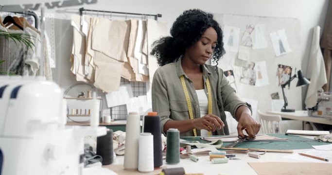 African American good looking young female tailor sitting at the table in her workshop studio and working over new clothes.