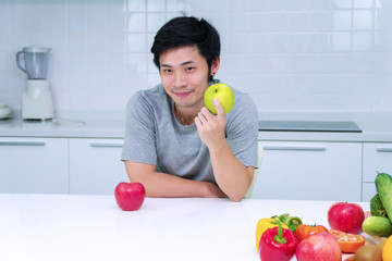 Healthy diet concept. Attractive smiling young man holding green fresh apple breakfast in his kitchen looking camera. Handsome male with vegetable and fruit on table at home.
