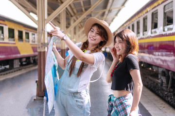 Cheerful two traveler young woman standing in the train station. Joyful tourist girl wear hat holding map in her hand, pointing finger with her friend or sister, talk about direction transit on trip.