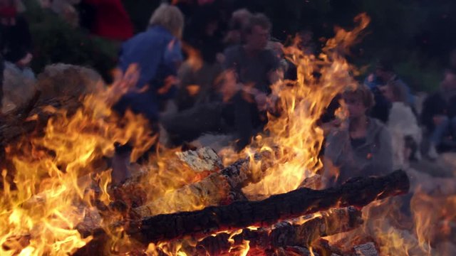 Closeup of Flames Blazing from a Pile of Wood with People in the Background