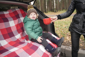 cute little kid boy sits in the trunk of a car on a red plaid and enjoys an autumn picnic alone