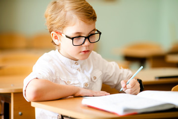Seven years old child with glasses writing his homework at school. Boy studing at table on class background