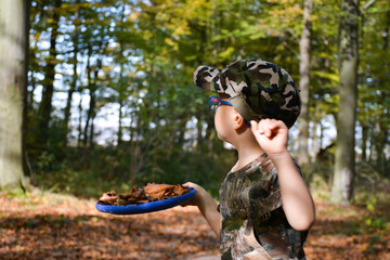 A child in camouflage clothes with a plastic plate in his hands collects autumn leaves