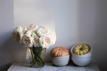 bouquet of white roses in a glass vase and pumpkins in a bowl on a white table as a decoration of the kitchen interior