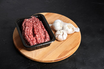 Fresh raw minced meat in tray and whole white garlic heads on round wooden cutting board on black stone background. Uncooked mincemeat. Ground beef and garlic bulbs