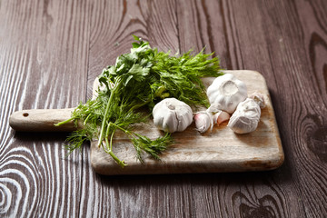 Cutting board with fresh seasonings on a wooden background. White garlic heads and cloves, green dill and parsley leaves. Condiments