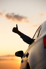 Man showing thumbs up/making Like / Ok sign with hand from car window with sunset sky, relaxing,...