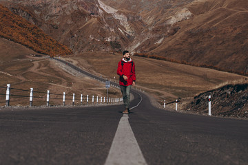 young attractive man with a beard dressed in a red jacket climbs the asphalt road on the slopes of the Caucasus Mountains against the backdrop of Mount Kazbek