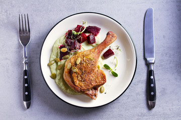 Roast duck leg with mashed green vegetables, beetroot, and pumpkin seeds on light gray background. Overhead image