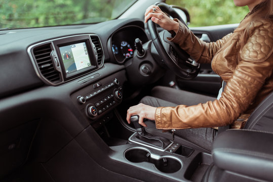 Close-up, woman driving a car includes a gearbox, reversing, starting to move in woods, leather jacket, automatic transmission. Touch screen navigation map app on monitor.