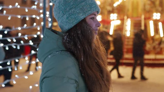 Smiling woman walking with sparkler in festive illumination tunnel on snowy night. Pretty girl in winter outfit flirt hanging at Christmas market. Garland lamps, carousel lights on background.