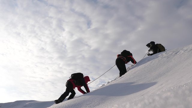 three Alpenists in winter climb rope on mountain. Travelers climb rope to  their victory through snow uphill in strong wind. tourists in winter work  together as team overcoming difficulties. Stock Photo