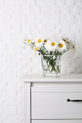 Beautiful tender chamomile flowers in vase on wooden commode near white textured wall