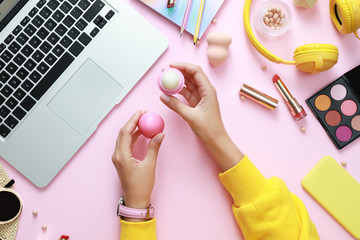 Young beauty blogger with lip balm near laptop and accessories on light pink background, top view