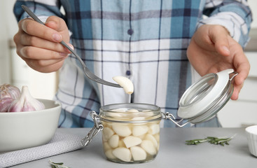Woman taking clove of pickled garlic from jar at grey table in kitchen, closeup