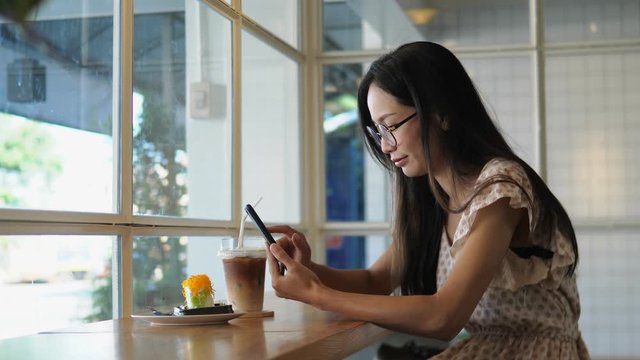 Young Asian woman using smartphone drinking coffee in cafe chatting with friends. Girl using cellphone in coffee shop.