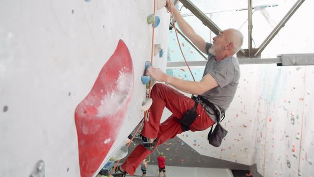 Aged slim bearded Caucasian climber wearing safety belay practicing climbing on climbing wall indoors