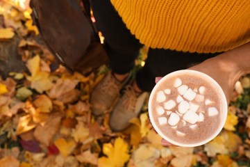 Woman holding cup of hot drink in park with fallen leaves, above view. Autumn season