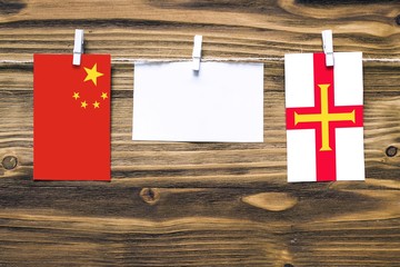 Hanging flags of China and Guernsey attached to rope with clothes pins with copy space on white note paper on wooden background.Diplomatic relations between countries.