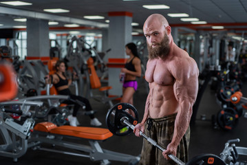 Fototapeta na wymiar Bald man with a beard in the gym. Muscular bodybuilder guy doing barbell exercises. Strong man with a naked torso. The young athlete is preparing for weightlifting competitions.