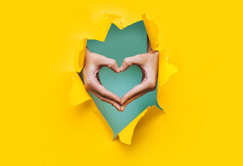 Female hands show a heart symbol through the torn holes of a yellow and green paper background....