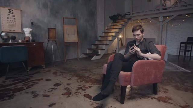 Attractive young man sitting in comfortable red armchair with smartphone in beautiful dark studio with decorative furniche. Stock footage. Professional photo and video shooting