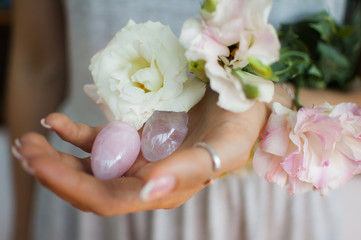 Female hands holding two yoni eggs for vumfit, imbuilding or meditation are made from pink quartz and transparent violet amethyst with white flowers indoors