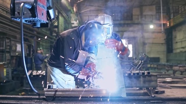 Tracking shot of unrecognizable worker in safety helmet and gloves helping trainee using welding tool on steel part at metal fabrication facility