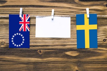 Hanging flags of Cook Islands and Sweden attached to rope with clothes pins with copy space on white note paper on wooden background.Diplomatic relations between countries.