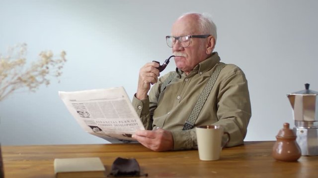 Dolly-in portrait shot of senior man with grey hair and mustache sitting at table and smoking pipe while reading newspaper, then looking at camera and smiling