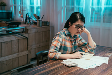 Fototapeta na wymiar Indoor shot of casually dressed young asian woman frowning reading at paper calculating family budget trying to save some money. lady wife in stressed and concentrated look in night kitchen.
