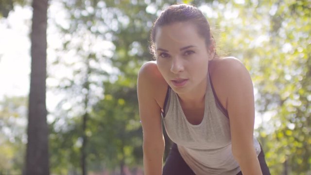 Young beautiful female athlete in sportswear standing with hands on knees, heavily breathing and looking at camera while resting after run in park on summer day