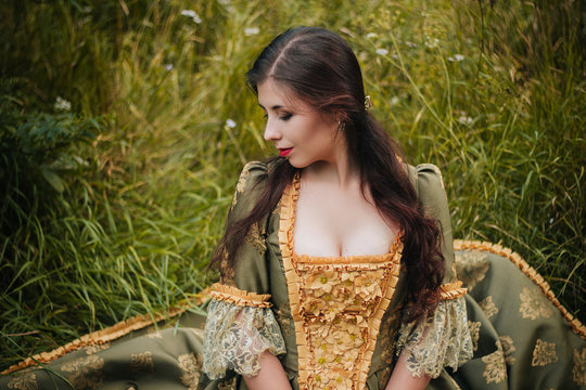 Young beautiful girl in a green rococo dress posing while sitting on the grass. Close-up portrait. A model with clean skin.