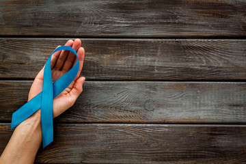 Blue ribbon in hands as symbol disease control on dark wooden background top view copy space