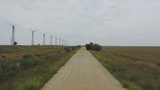 Aerial view glight above dirt road with spinning wind turbines and field