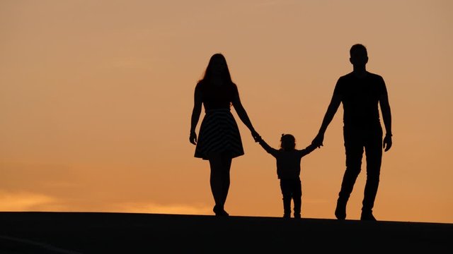 Young family concept. Silhouettes of mother, little daughter and father walking together. Man and woman holding hands of small baby, toddle between, just started to walk