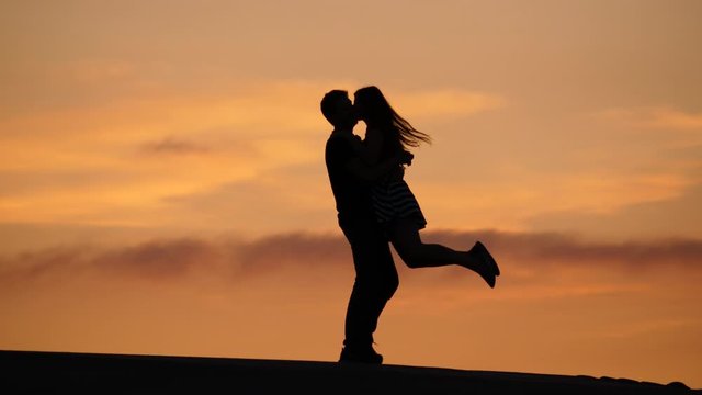 Happy man embrace woman, hold her tight and turn around, silhouetted figures against sunset sky. Beautiful couple celebrate or have fun, hug and spin