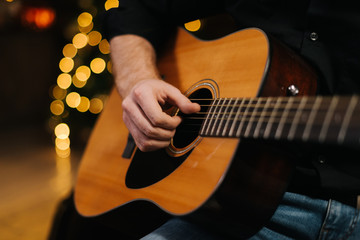 Man plays guitar close-up. Against the background of a decorated Christmas tree with a bokeh effect. - obrazy, fototapety, plakaty