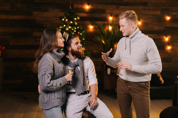 Guy talks to couple in celebrating new years eve party in festively decorated house holding glasses of champagne in hand. Christmas tree with garland and festive illumination in background.