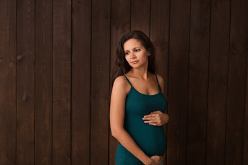 Pregnant woman in jeans shorts and green top holds hands on belly on a dark brown background. Pregnancy, maternity, preparation and expectation concept. Beautiful tender mood photo of pregnancy