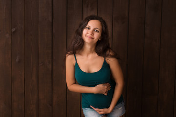 Pregnant woman in jeans shorts and green top holds hands on belly on a dark brown background. Pregnancy, maternity, preparation and expectation concept. Beautiful tender mood photo of pregnancy