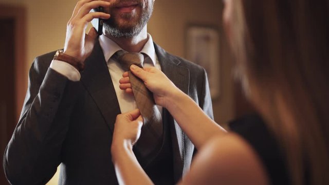 Young man in suit talking on a mobile phone, woman ties a tie to a man, business trip, hotel interior.