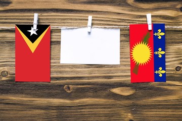 Hanging flags of East Timor and Guadeloupe attached to rope with clothes pins with copy space on white note paper on wooden background.Diplomatic relations between countries.