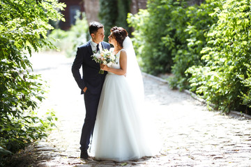 The bride and groom walk together in the park. Charming bride in a white dress, the groom is dressed in a dark elegant suit. Pretty bride and stylish groom. Wedding day. Marriage.
