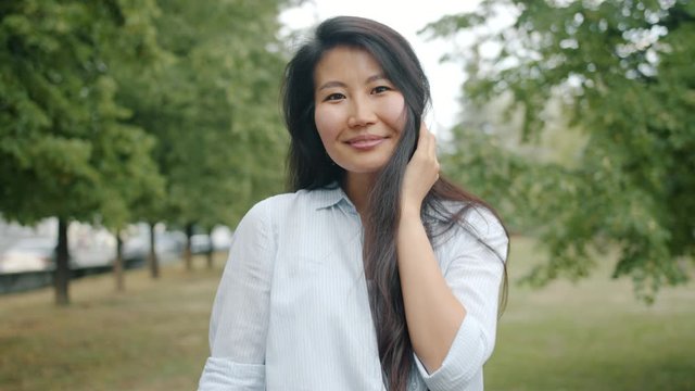Cheerful Asian lady student is flirting in park touching hair smiling looking at camera alone with nature lawn and trees in background. People and emotions concept.