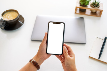 Top view mockup image of hands holding mobile phone with blank screen and laptop computer on the table in office