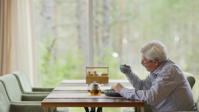Side view medium out shot of senior man with grey hair and in eyeglasses sitting alone at cafe table, drinking tea and having dinner