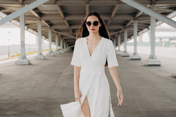 Fototapeta premium young woman in the city. Portrait of a young woman in sunglasses. The girl in the white dress. Walk around the city. Long-haired brunette. Female portrait. Beauty and fashion. Gray background. Bridges